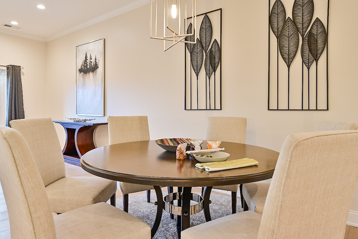 A dining room with a round table, beige chairs, wall art of leaves, a center bowl with items, and a console table with a landscape picture.