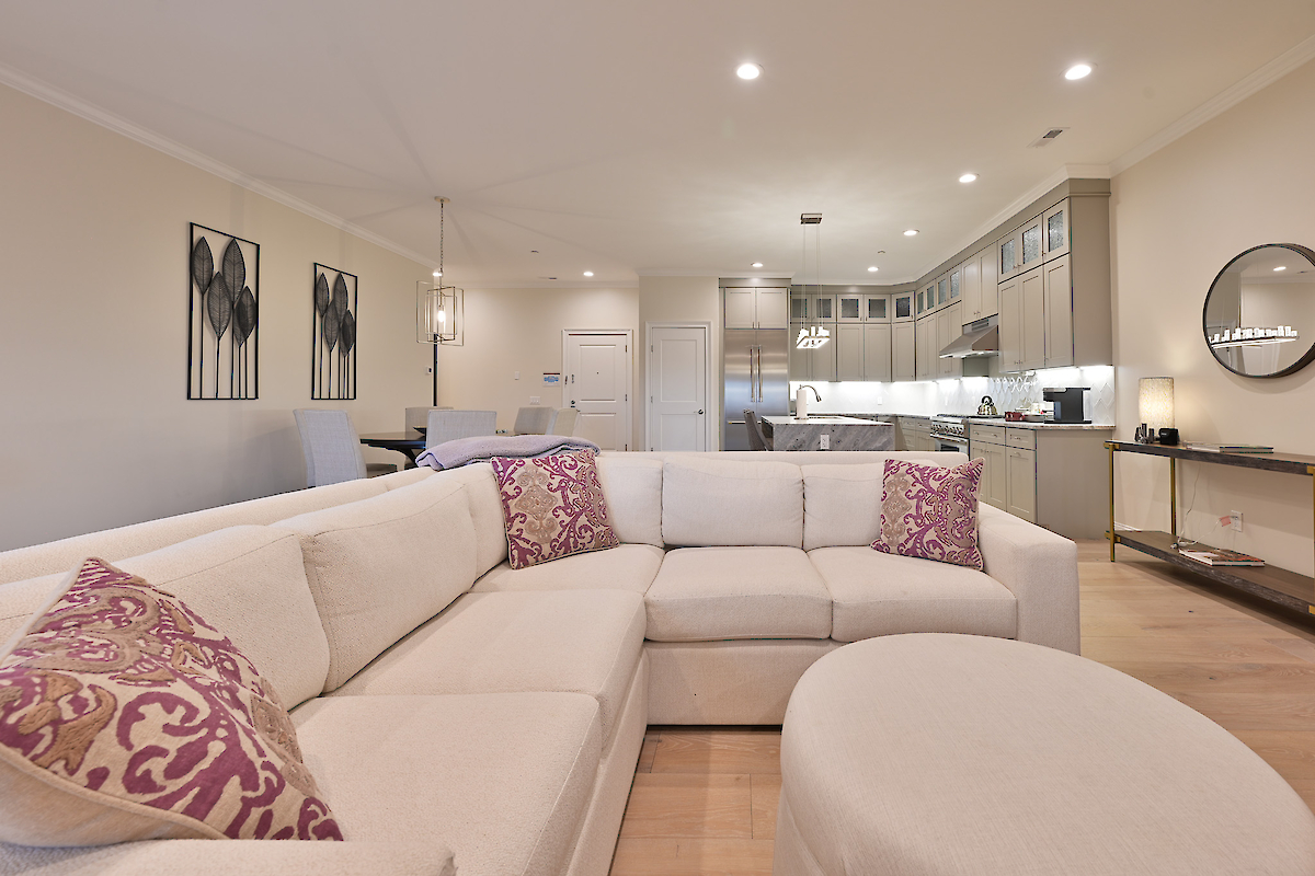 A modern living room with a beige sectional sofa featuring purple cushions, open to a kitchen and dining area under bright lighting.