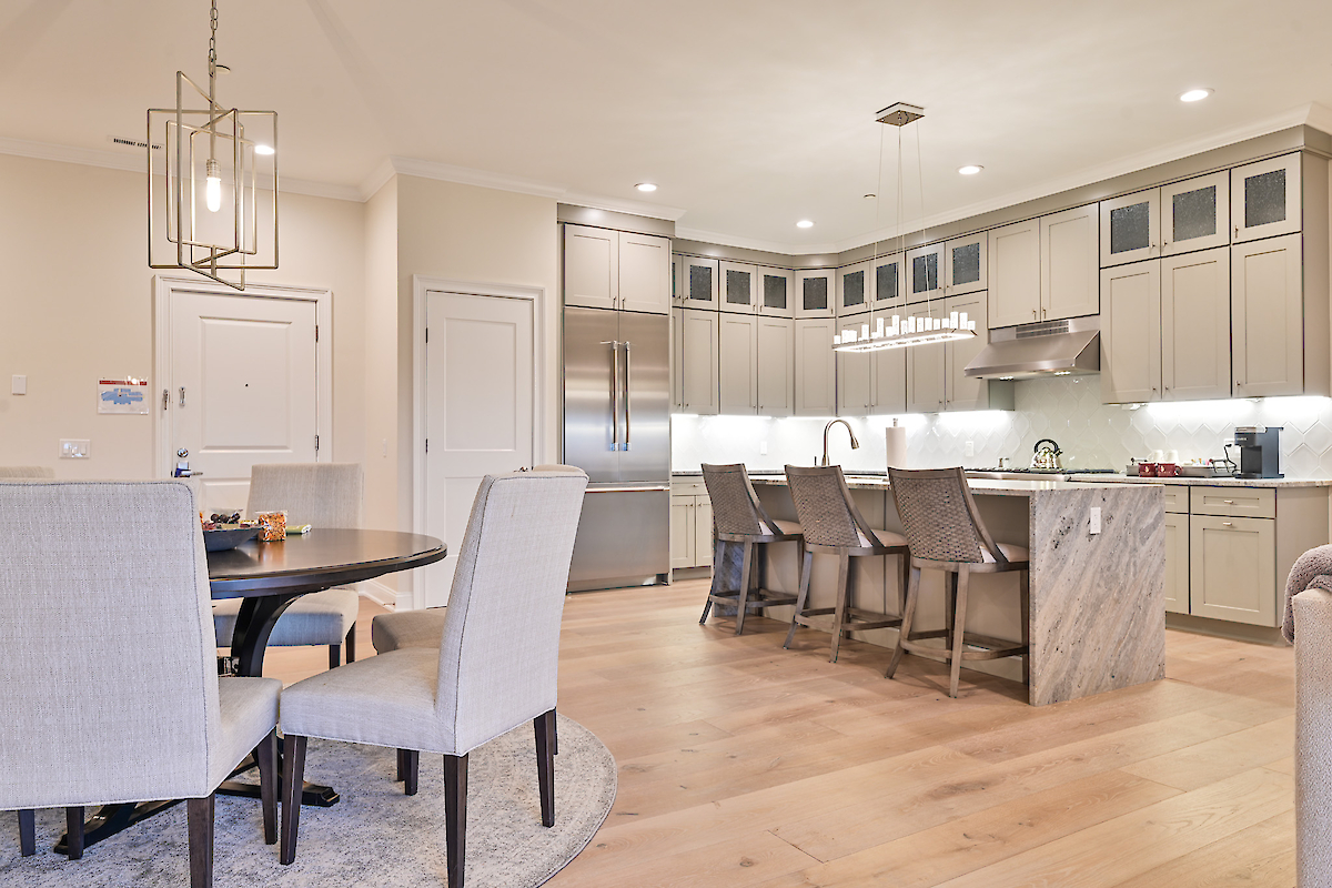A modern kitchen and dining area with a round table, cushioned chairs, a kitchen island with stools, stainless steel appliances, and pendant lighting.