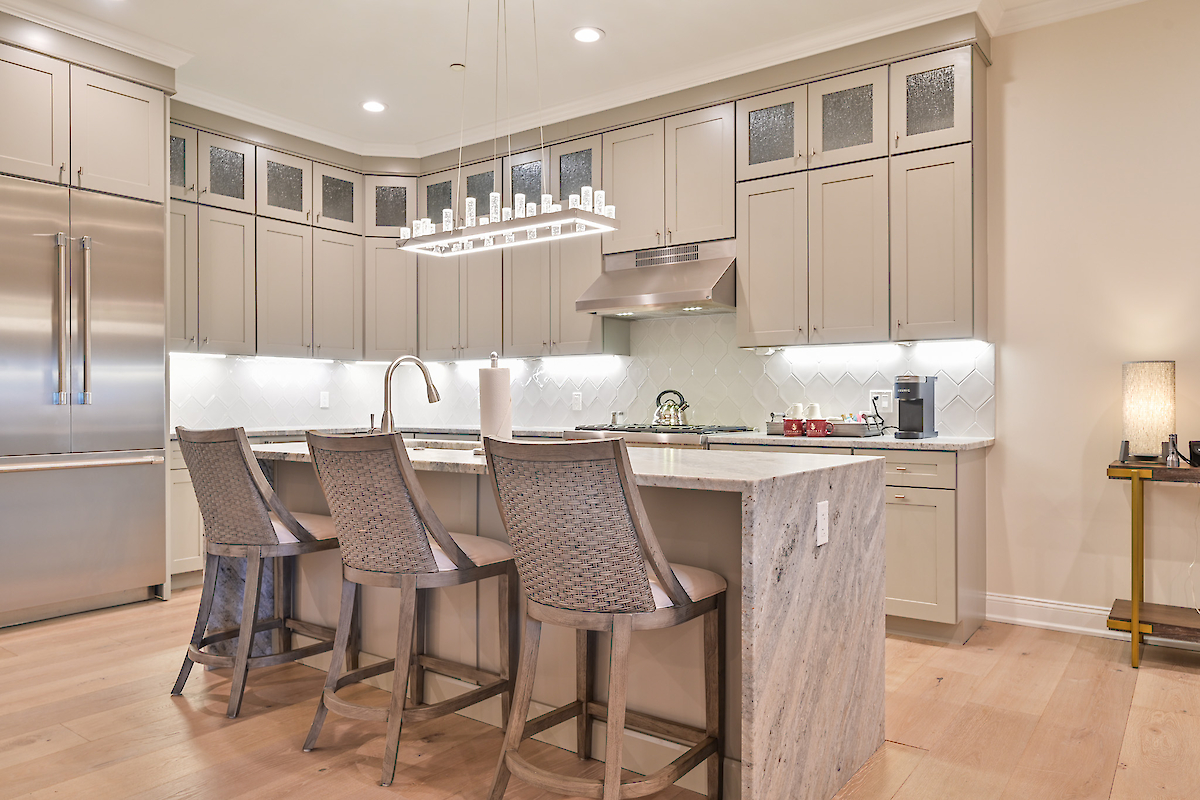A modern kitchen with an island, four chairs, stainless steel appliances, beige cabinets, and pendant lighting over the island.