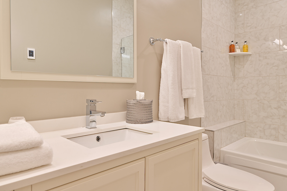 A tidy, modern bathroom with a white countertop, sink, towels, toiletries, a toilet, and a bathtub with a tiled wall.