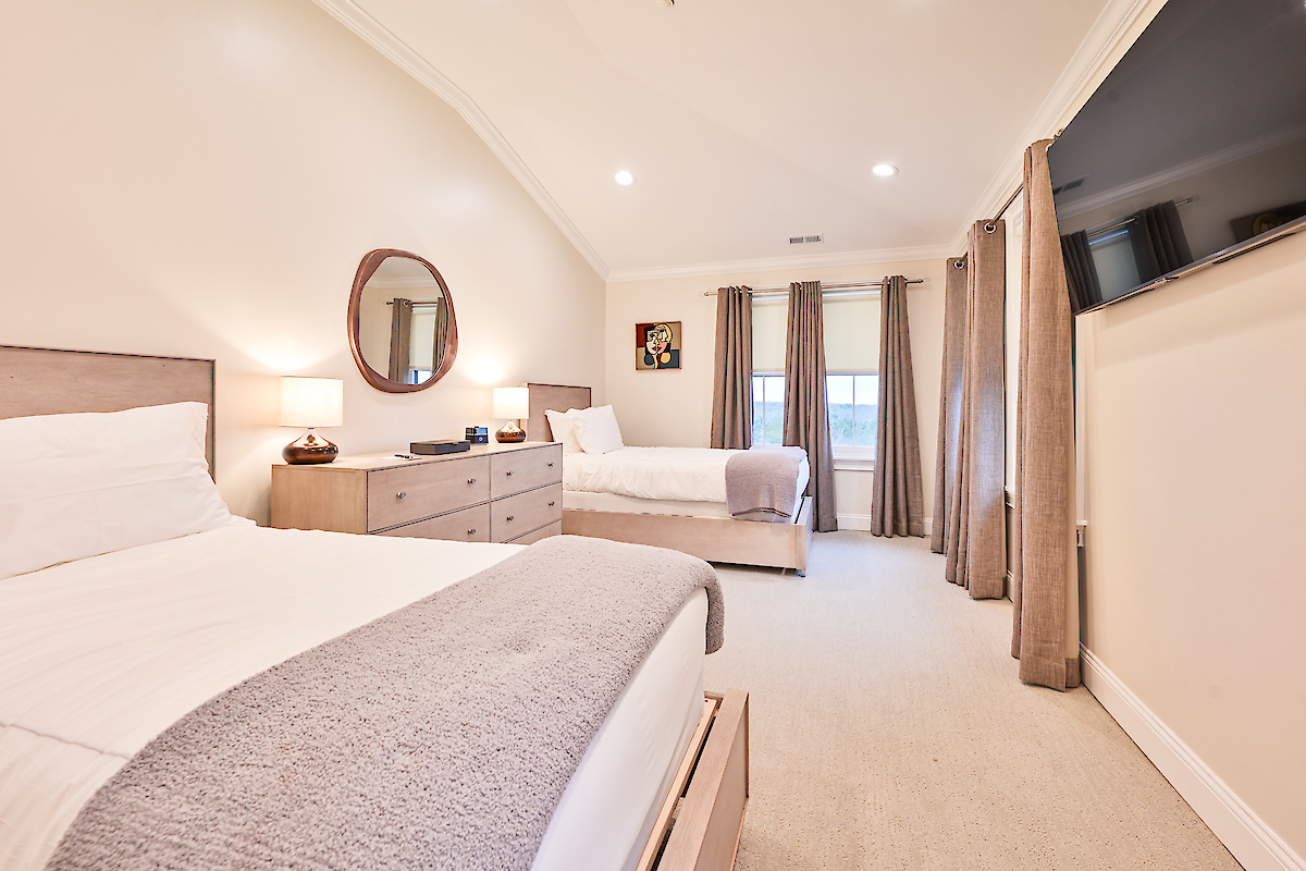 A well-lit bedroom features two beds, a dresser with a round mirror, lamps, a wall-mounted TV, and large windows with curtains, ending the sentence.