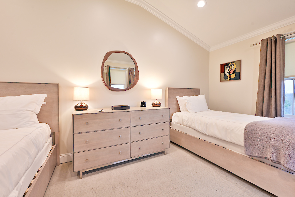 A bedroom with two single beds, a wooden dresser with two lamps, a round mirror, and a wall painting. The walls are beige in color.