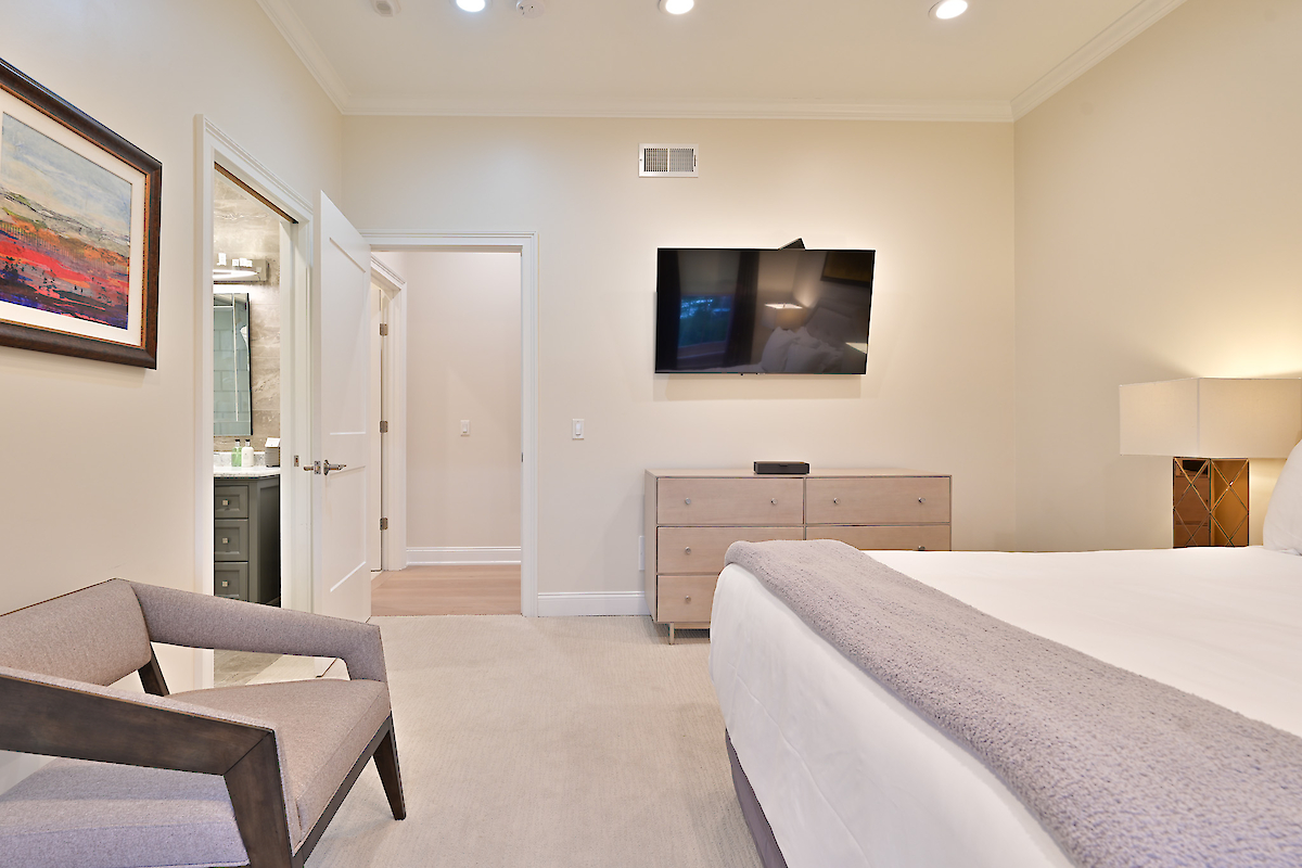 This image shows a modern bedroom with a wall-mounted TV, a dresser, a bed, a chair, a lamp, framed artwork, and a view into a bathroom.