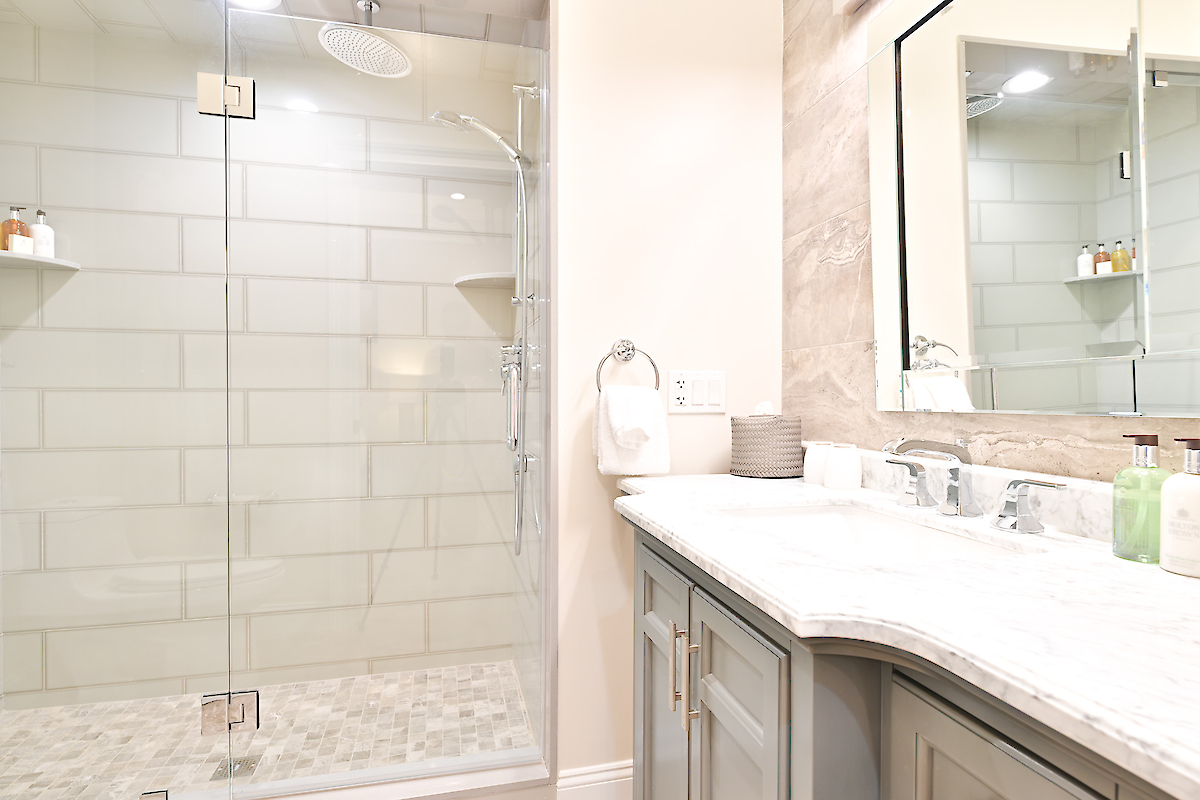 This image shows a modern bathroom with a glass-enclosed shower, a marble countertop with double sinks, a mirror, and various toiletries.