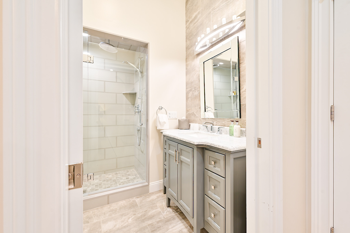 A bathroom with a glass-enclosed shower, a vanity with dual sinks, a large mirror, and overhead lighting. Beige walls and tiles.