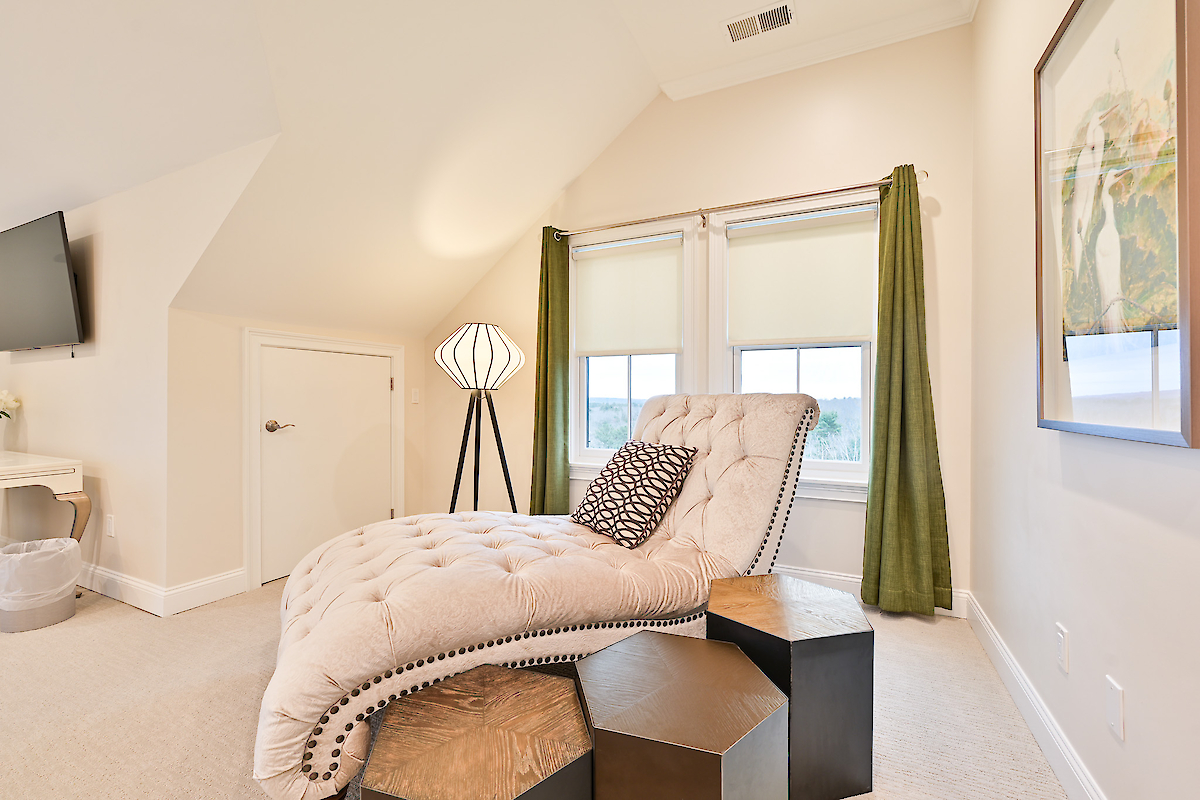 A well-lit room with a chaise lounge, patterned pillow, tripod lamp, two windows with green curtains, wall art, TV, and a side table.