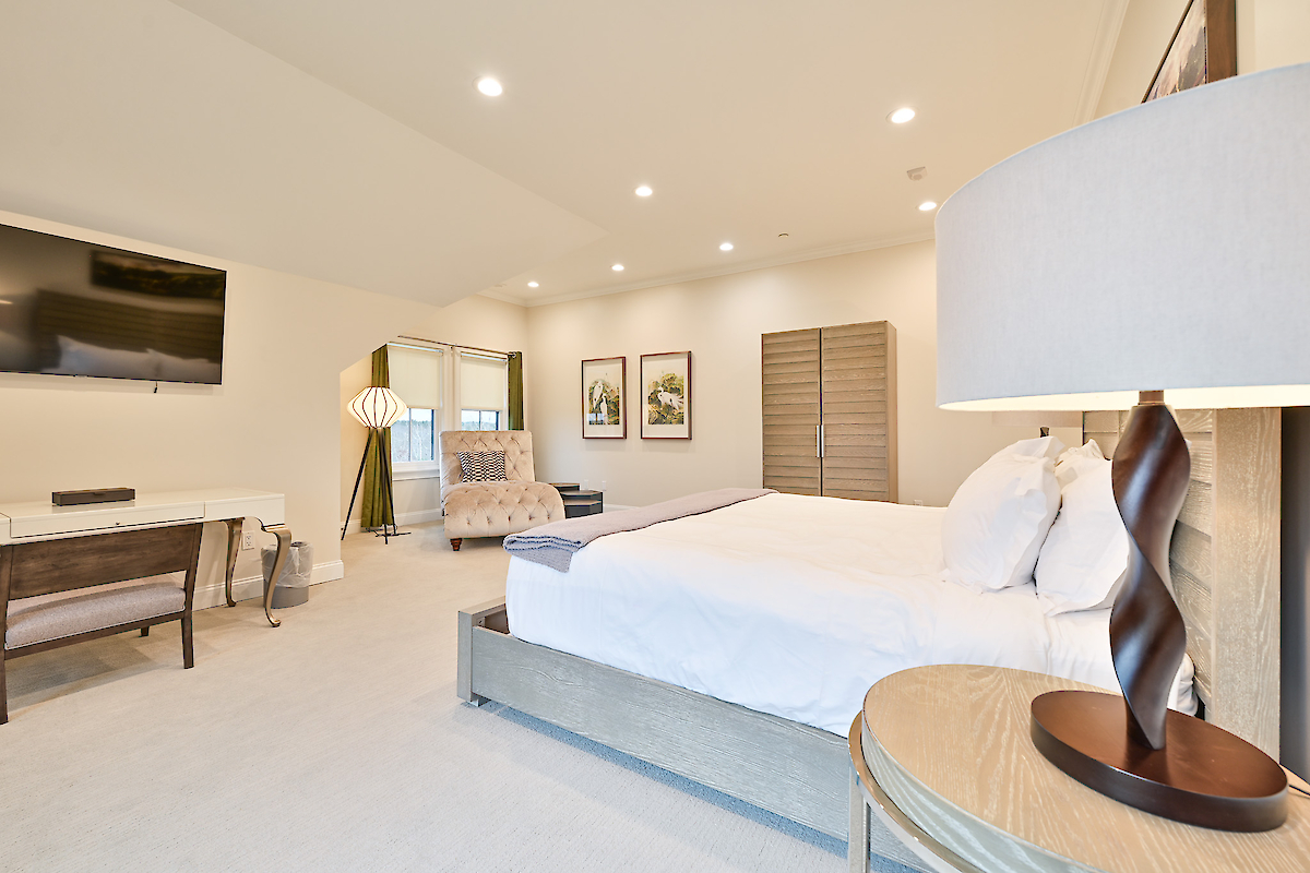 A modern bedroom with a large bed, desk, chair, TV, lamps, and wall art, featuring neutral colors and ample lighting.