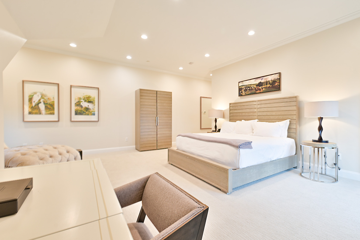 A modern bedroom with a large bed, desk, chair, and two lamps. Wall art and a wooden cabinet decorate the room, providing a cozy ambiance.