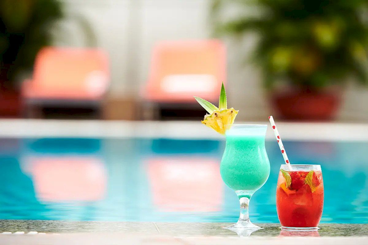 Two colorful cocktails with garnishes sit poolside, with lounge chairs and greenery in the blurry background.