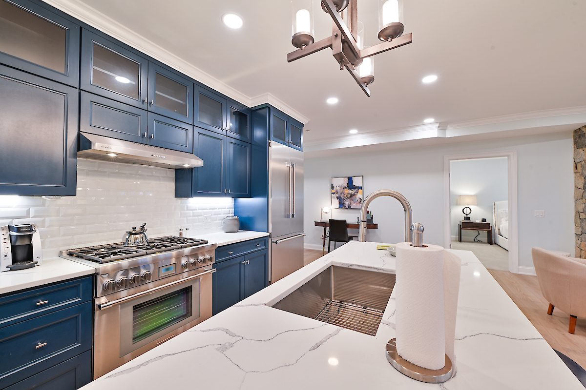 A modern kitchen featuring blue cabinets, a marble countertop with a sink, a stove, and an adjacent room in the background.