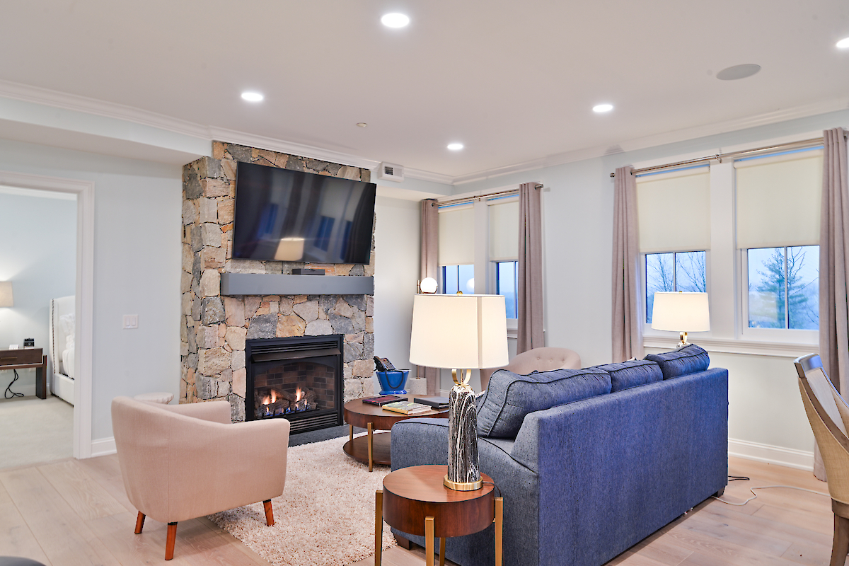 A cozy living room with a blue couch, armchair, stone fireplace, wall-mounted TV, side table, and lamps. Windowed room with natural light.