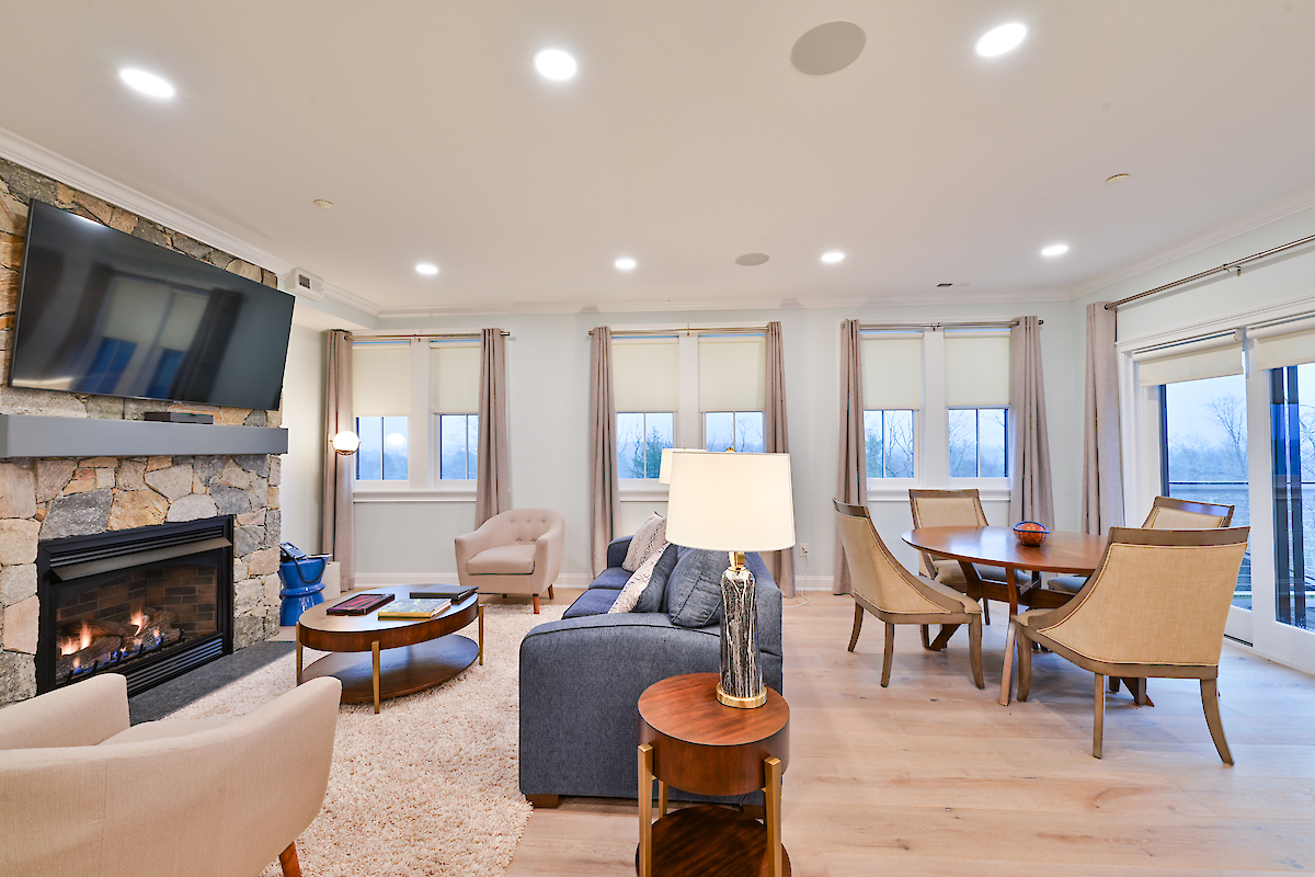 A modern living room with a fireplace, mounted TV, sofas, chairs, a dining table, and large windows, featuring wooden flooring and recessed lighting.