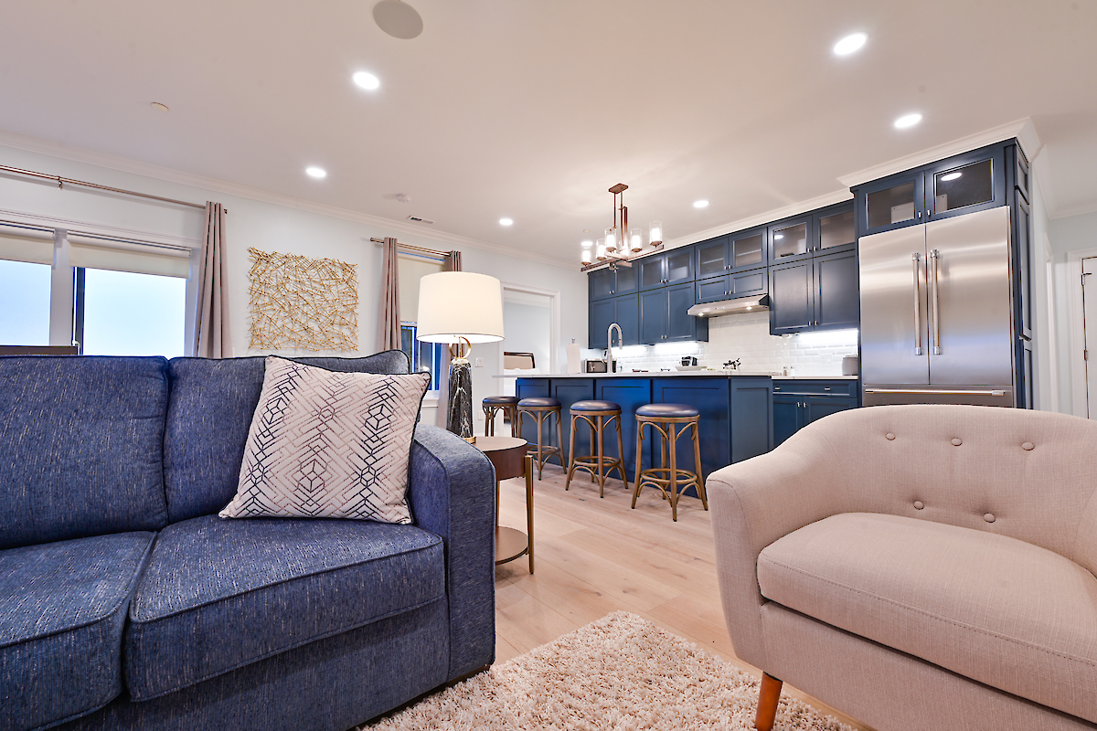 A modern living area with a blue sofa, beige chair, and open kitchen featuring dark blue cabinets, stainless steel appliances, and high stools at the counter.