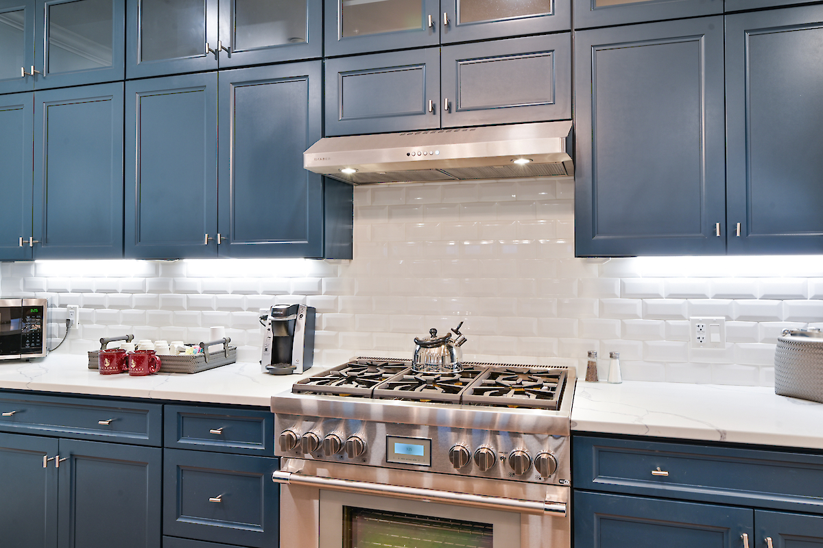 A modern kitchen with blue cabinets, stainless steel stove, white tiled backsplash, countertop appliances, and under-cabinet lighting.