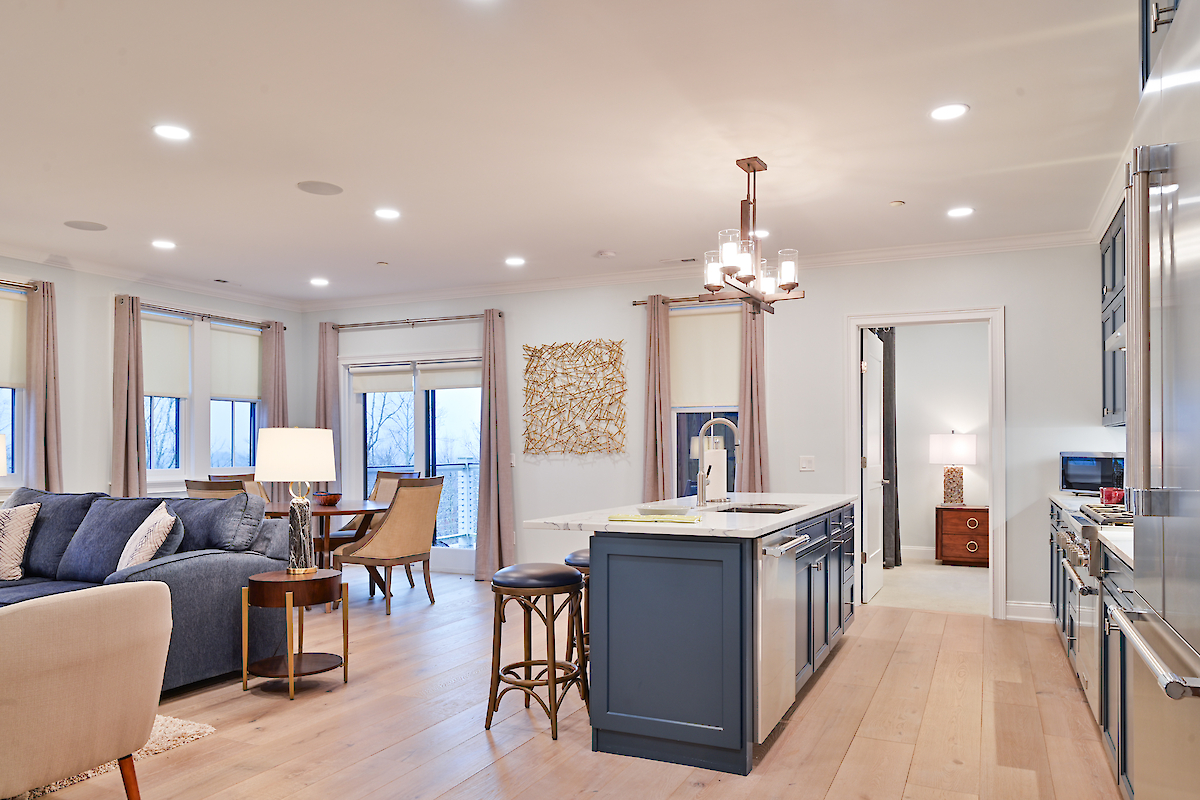 The image shows a modern open-concept living area with a kitchen island, seating area, and dining table, decorated in neutral tones with ample lighting.