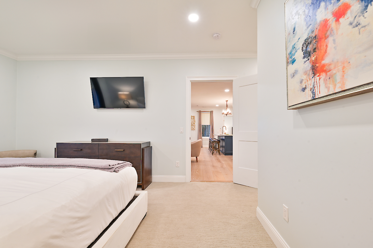 A bedroom with a bed, wall-mounted TV, dresser, and abstract painting, leading to an adjacent kitchen or dining area through an open door.