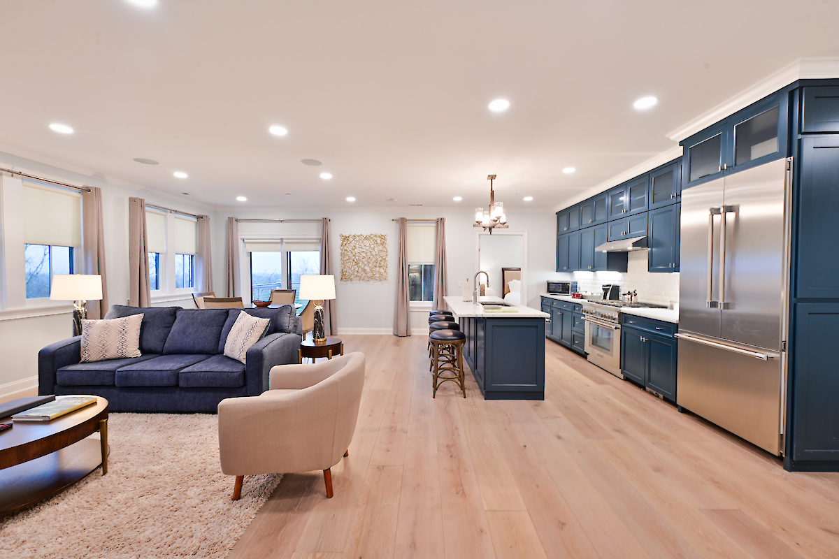A modern open-concept living room and kitchen with blue cabinetry, stainless steel appliances, a sofa, armchair, and island with bar stools.