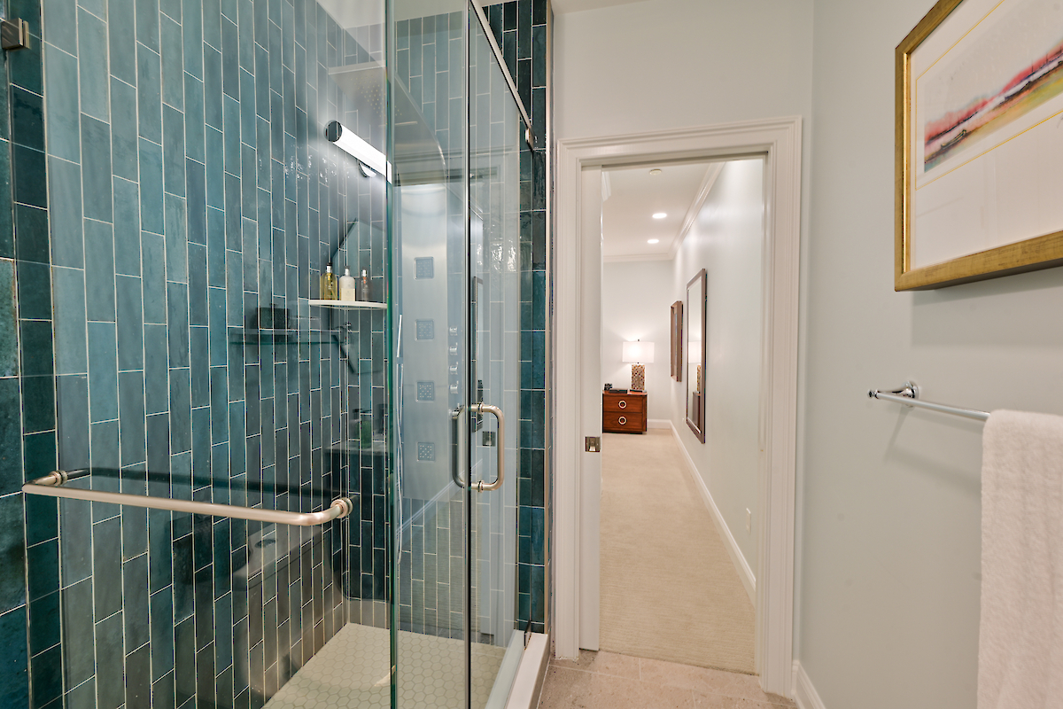 A modern bathroom with a glass shower enclosure, blue tiles, towel rack, and a view into an adjoining hallway featuring a dresser.