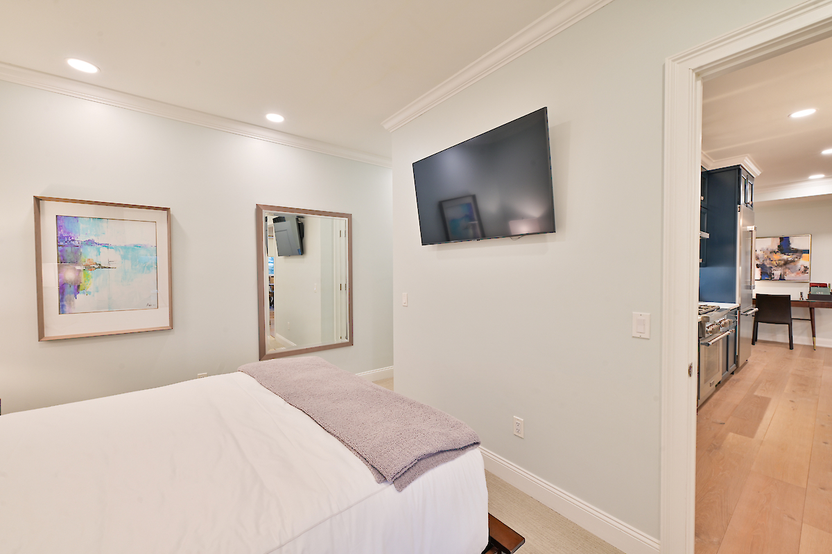 The image shows a bedroom with a bed, a wall-mounted TV, a large mirror, a framed picture, and an open doorway leading to another room.