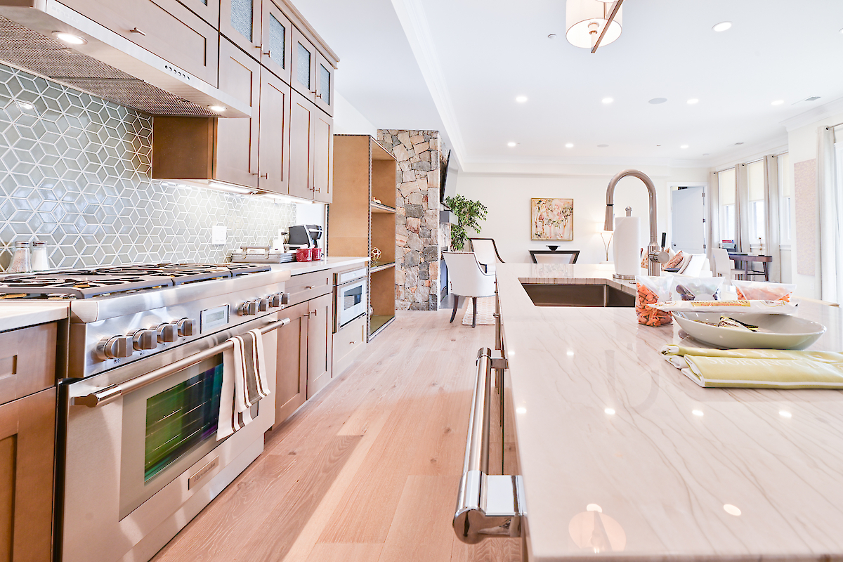 Modern kitchen with wooden cabinets, a tiled backsplash, large stovetop, island with sink, and an open floor plan leading to dining area.