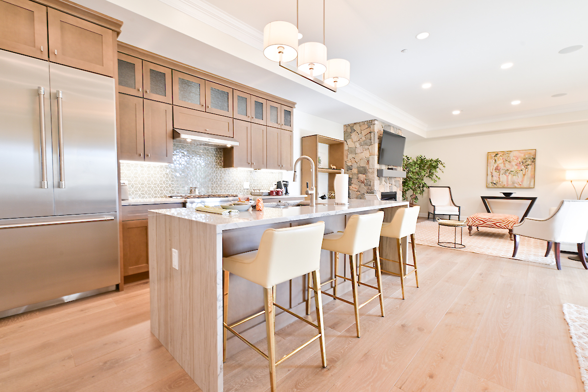 A modern kitchen with a large island, bar stools, stainless steel appliances, and light wooden cabinets, ending with a cozy living area.