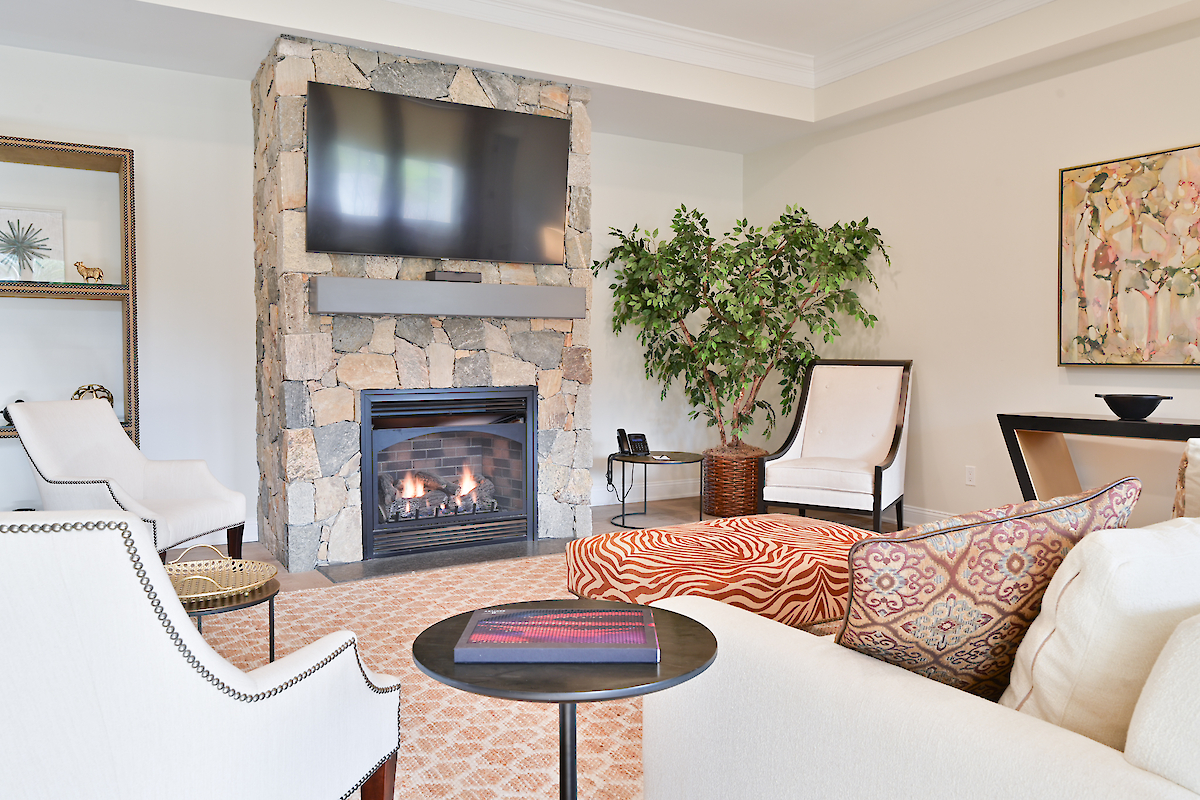 A cozy living room with a stone fireplace, TV, white chairs, a plant, a patterned ottoman, and a coffee table. Ends the sentence.