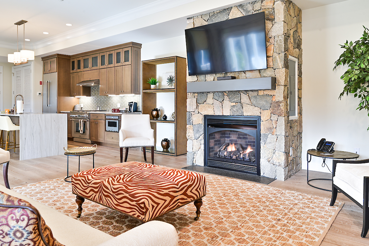 A modern living room with a stone fireplace, television, kitchen area, and patterned ottoman in front of sofas.