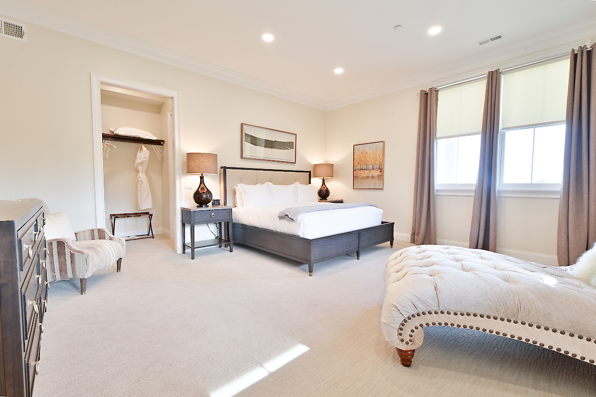 A well-lit bedroom with a large bed, chaise lounge, two windows, dark nightstands, lamps, and artwork. There's also a closet with clothes.