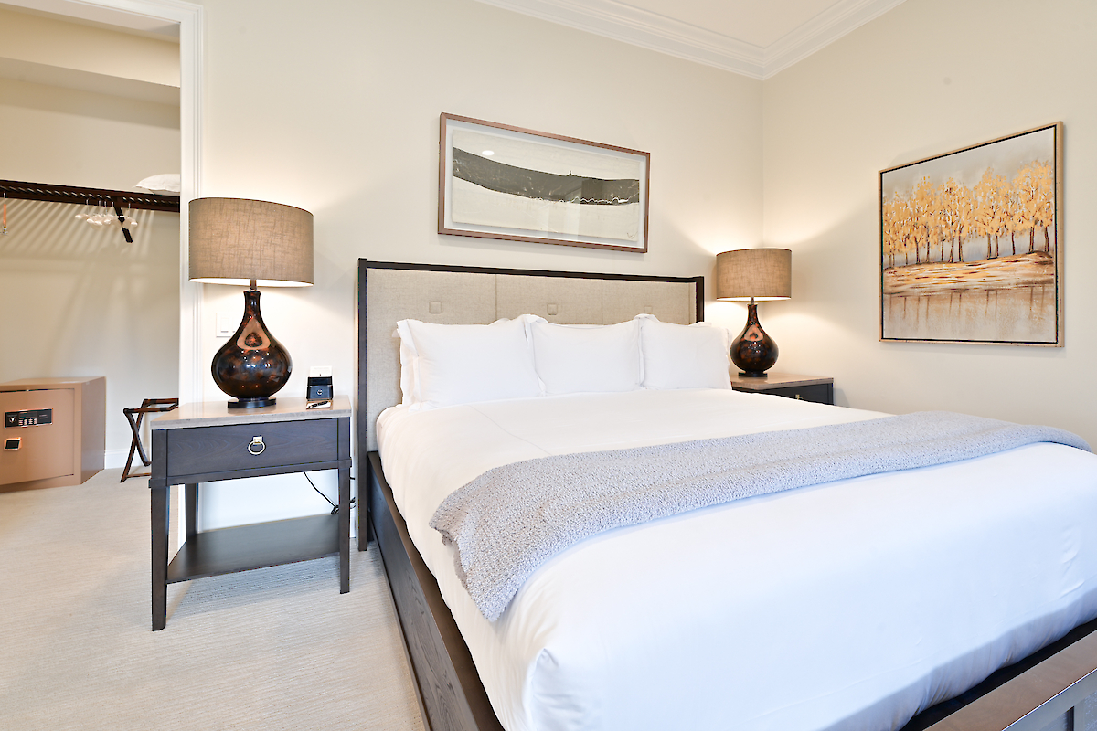 A bedroom with a neatly made bed, two bedside tables with lamps, artwork on the walls, and an open walk-in closet.