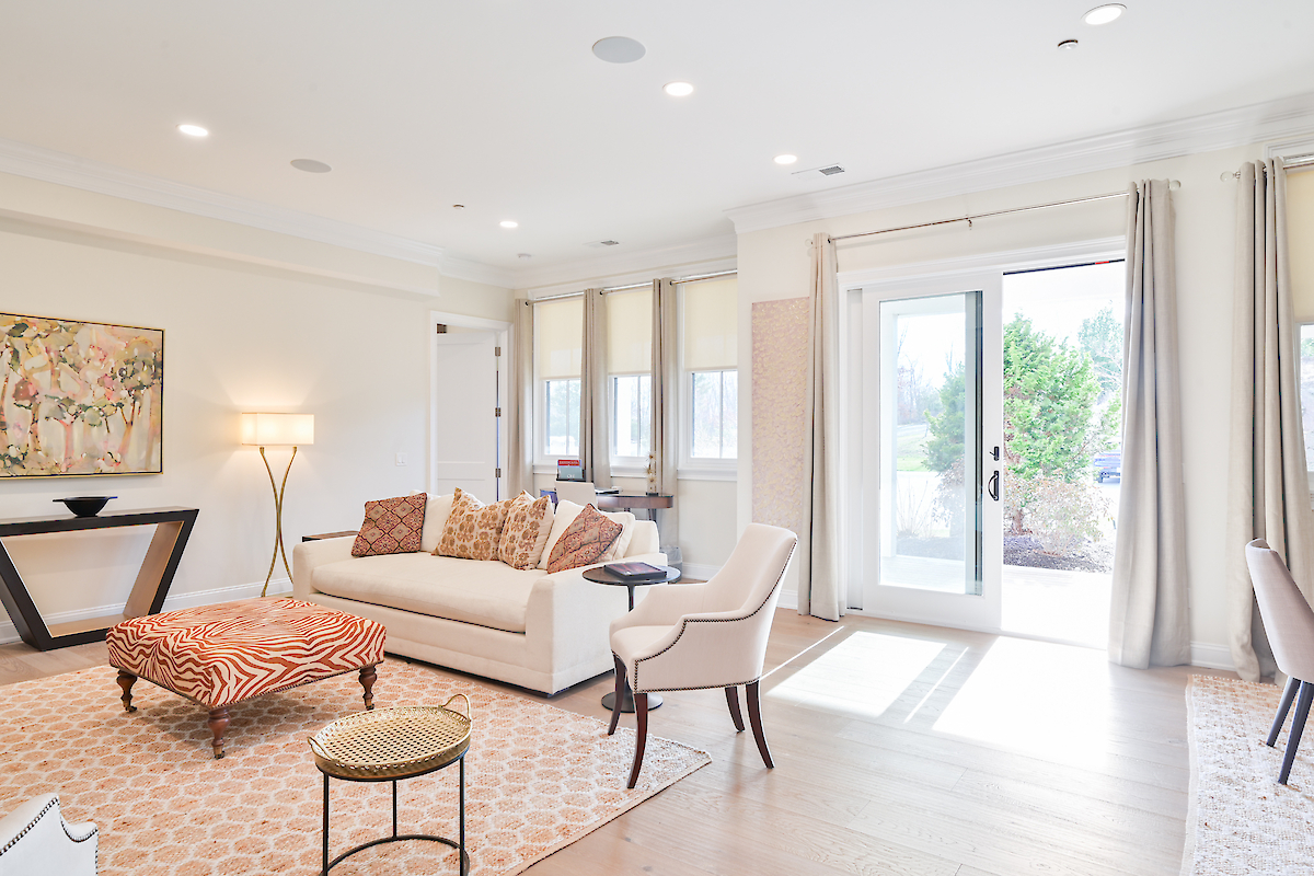 A bright living room with a beige sofa, patterned ottoman, art on the wall, floor lamp, and open French doors leading to the outside.