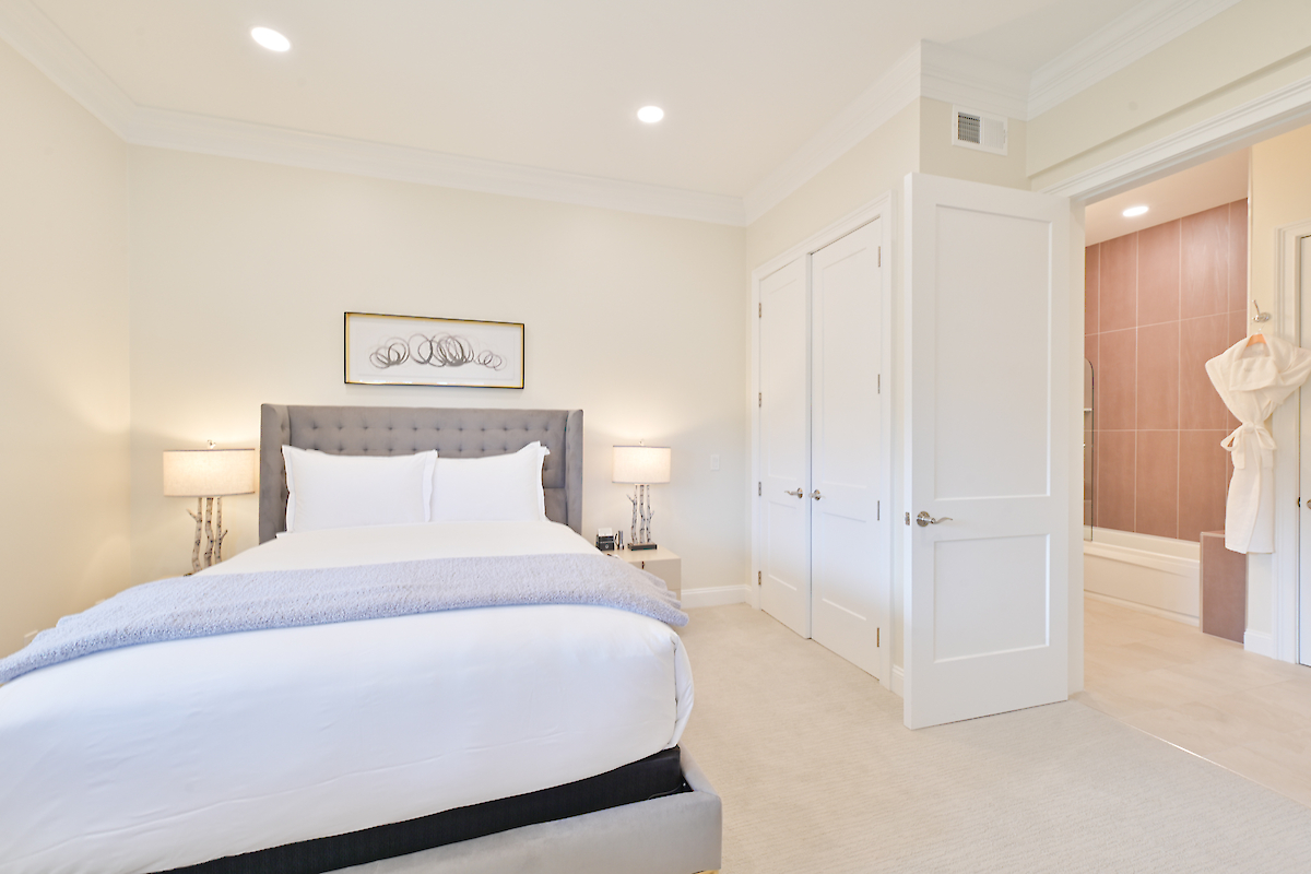 A neatly arranged bedroom features a bed, two side tables with lamps, a painting above the headboard, and an adjacent bathroom.