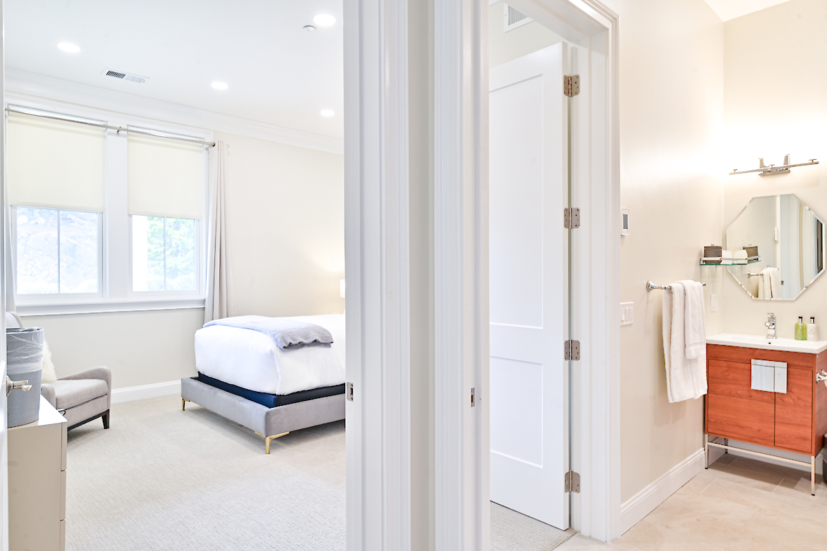 The image shows a bright bedroom with a bed, chair, and dresser, adjacent to a bathroom with a vanity, mirror, and towels on the rack.