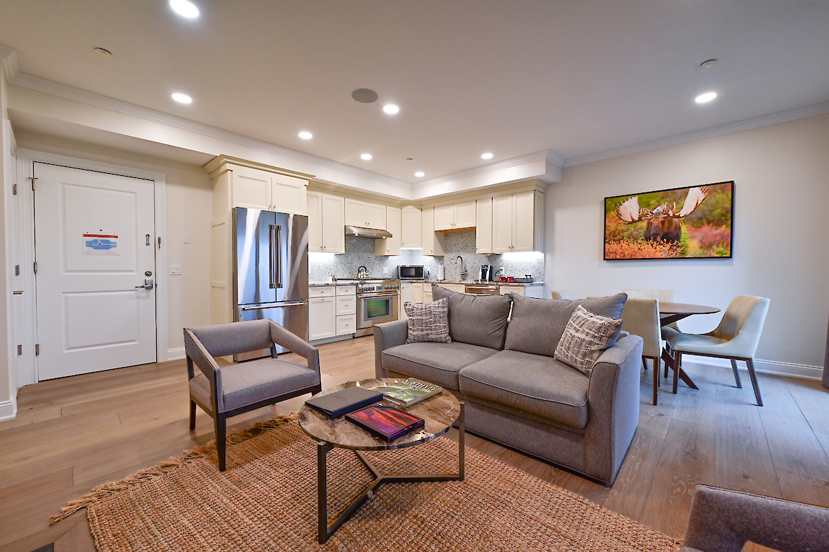 A modern open-plan living room and kitchen with a gray sofa, armchair, coffee table, dining area, and contemporary decor, including a wall painting.