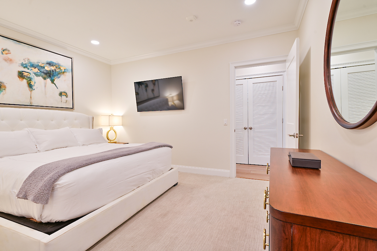 A modern bedroom features a large bed with white linens, a wall-mounted TV, a wooden dresser with a mirror, and a double-door closet.