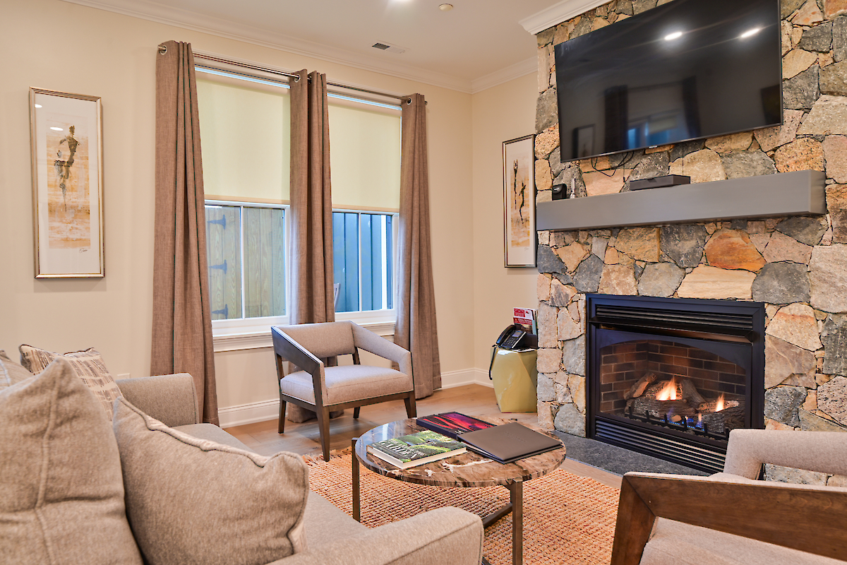 A cozy living room with a stone fireplace, mounted TV, two windows with curtains, and contemporary furniture, including a coffee table and chairs.