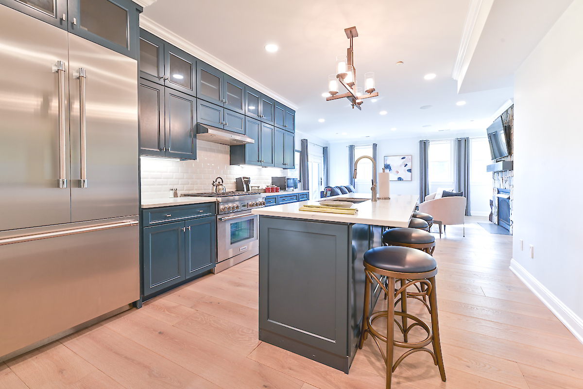 Modern kitchen with blue cabinets, stainless steel appliances, a central island with stools, and light wood flooring extending to a living area.