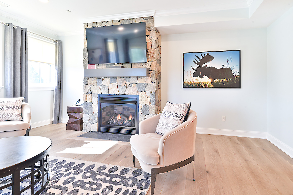A modern living room features a stone fireplace, flat-screen TV, moose artwork, beige chairs, patterned rug, and large windows.