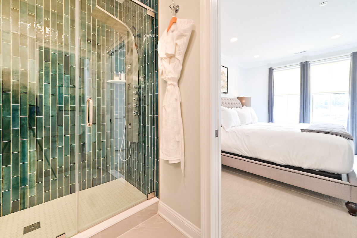 This image shows a shower with green tiles, a white bathrobe hanging on a hook, and a bedroom with a neatly made bed and curtains.