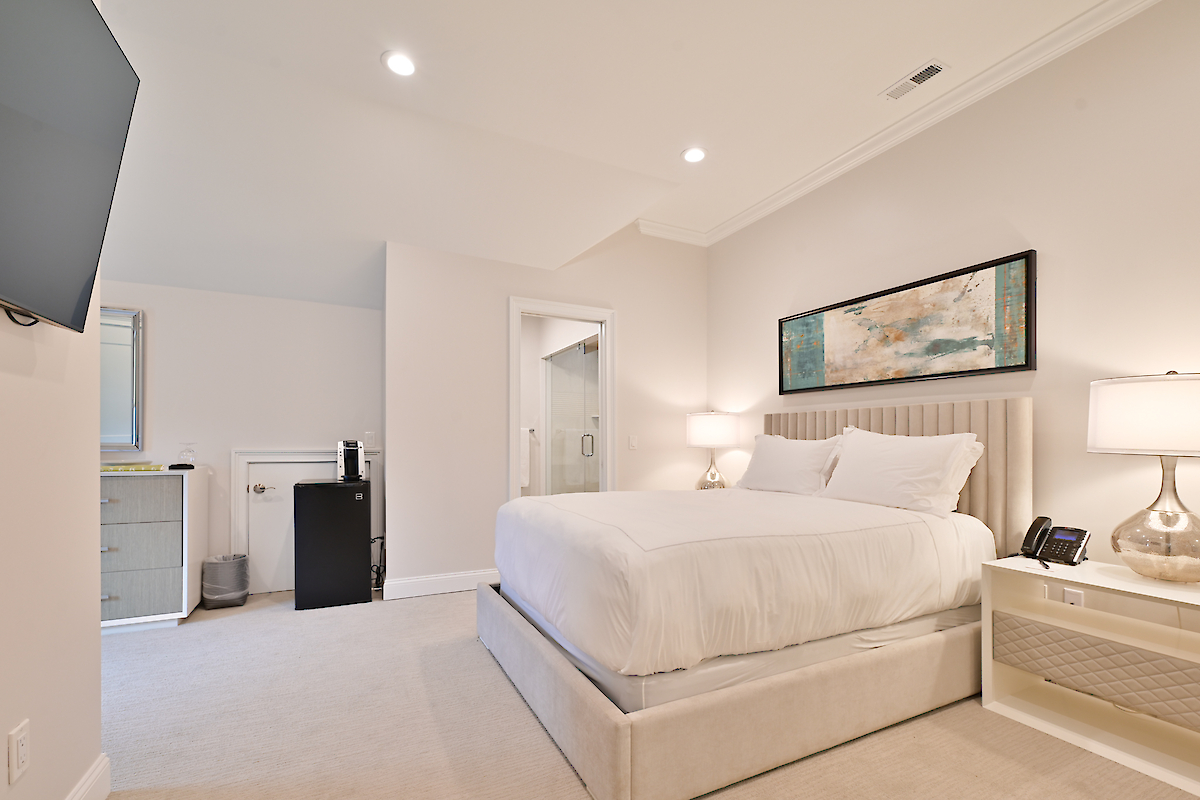 A modern bedroom with a neatly made bed, nightstands with lamps, a wall-mounted TV, dresser, and a mini-fridge, leading into a bathroom.