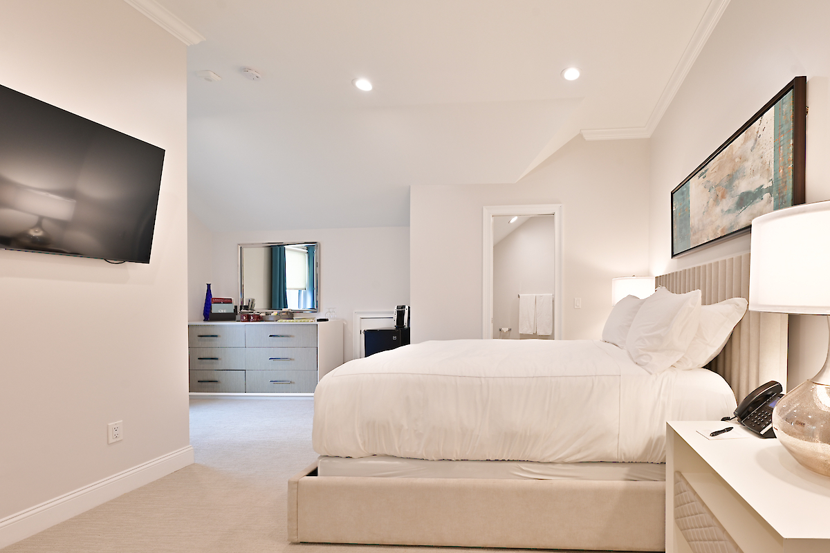 A modern bedroom with a wall-mounted TV, white bed, bedside table, chair, dresser, lamp, artwork on the wall, and an en-suite bathroom.