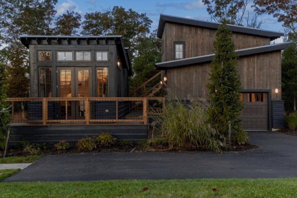 The image shows a modern wooden house with large windows, a front deck, an attached garage, surrounded by trees and a paved driveway.