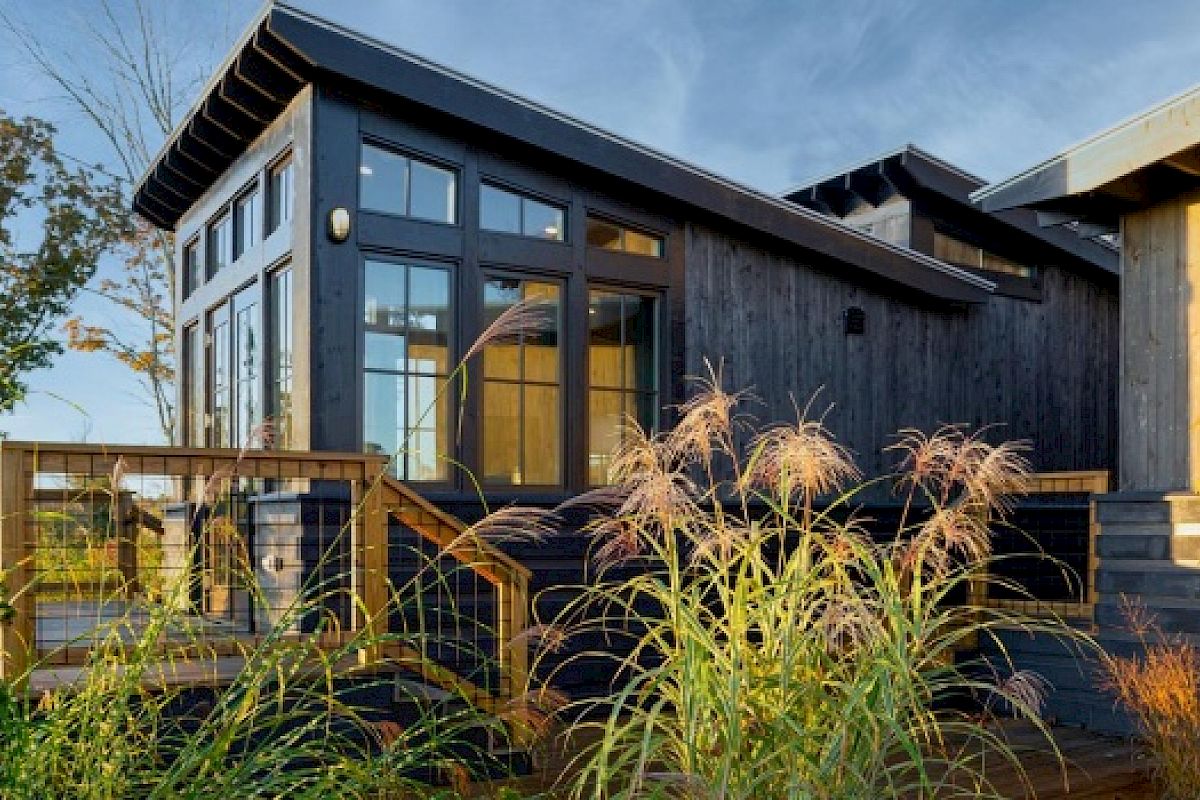 This image features a modern wooden house with large windows, a wooden deck, and lush grass in the foreground under a clear sky.