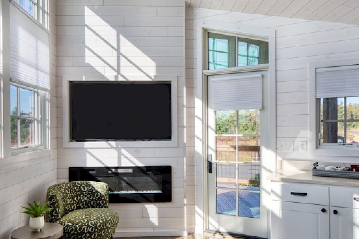 A bright room with large windows, a wall-mounted TV, a green patterned chair, a small plant, and a desk with drawers and books.