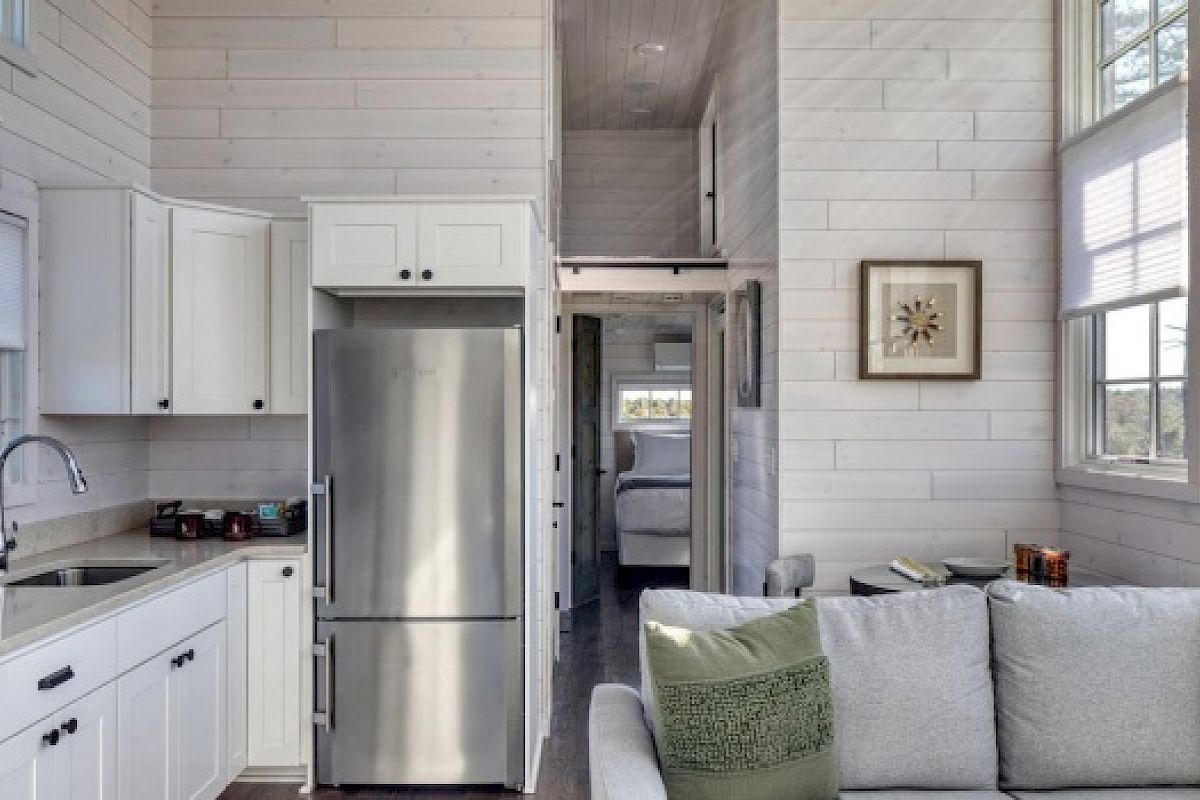 This image shows a modern tiny home interior with a kitchen on the left, a living area on the right, and a hallway leading to a bedroom.