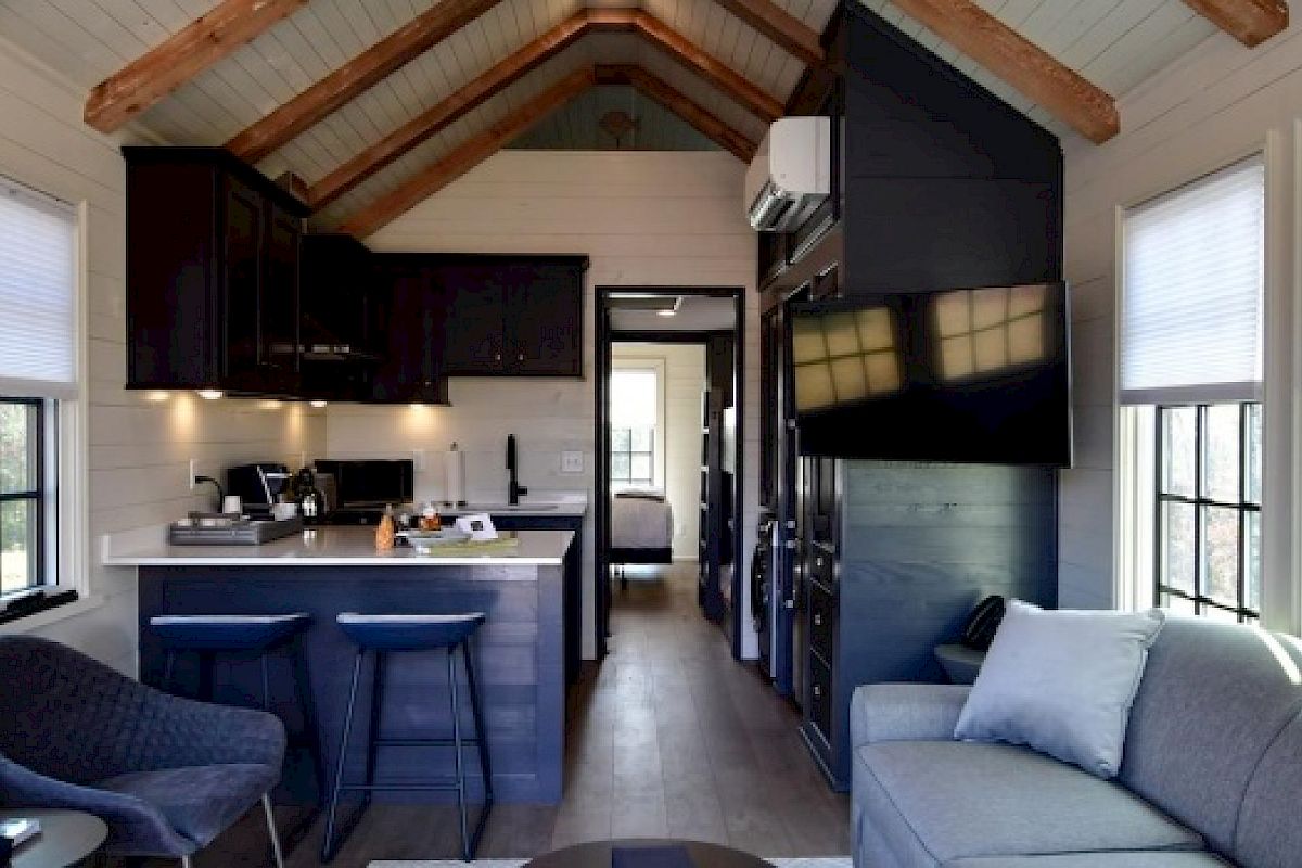 A cozy tiny house interior with a living area, kitchen, and bar stools, featuring a sofa, flat-screen TV, and loft-style wooden ceiling beams.