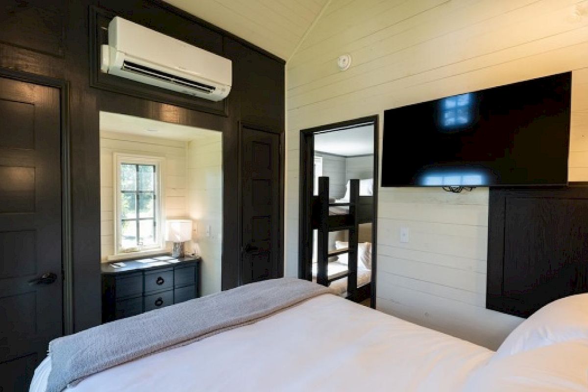 A cozy bedroom features a bed, a wall-mounted TV, an AC unit, and a small desk area by a window. Dark wooden accents complement white walls.
