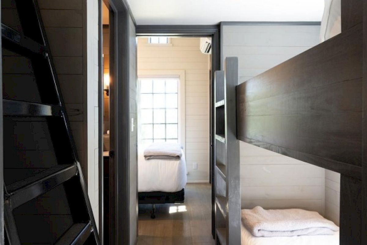 The image shows a narrow hallway with a bunk bed on the right, a ladder on the left, and a view into a bedroom with a window at the far end.
