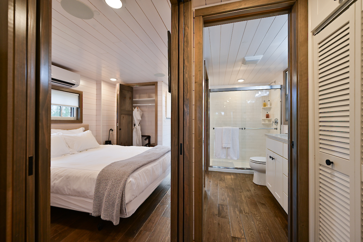 This image shows a neatly arranged bedroom with a white bed and a view into an adjacent bathroom. The room has wooden floors and white walls.