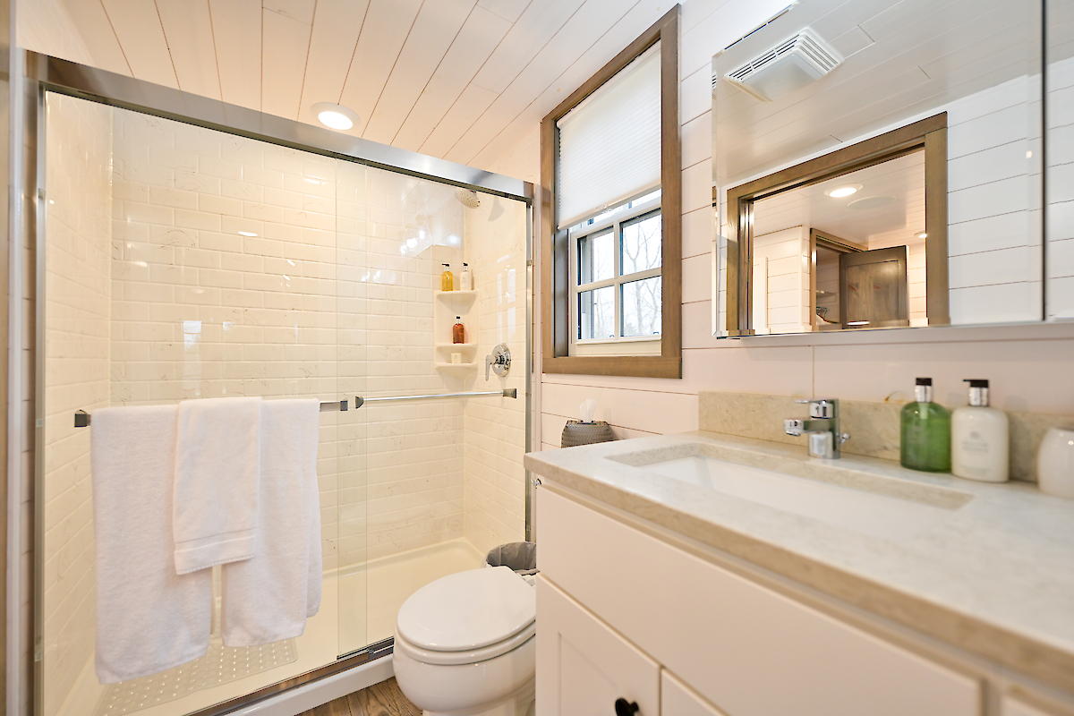 A modern bathroom with a glass-enclosed shower, sink with a mirror, and various toiletries on the countertop. A window adds natural light.